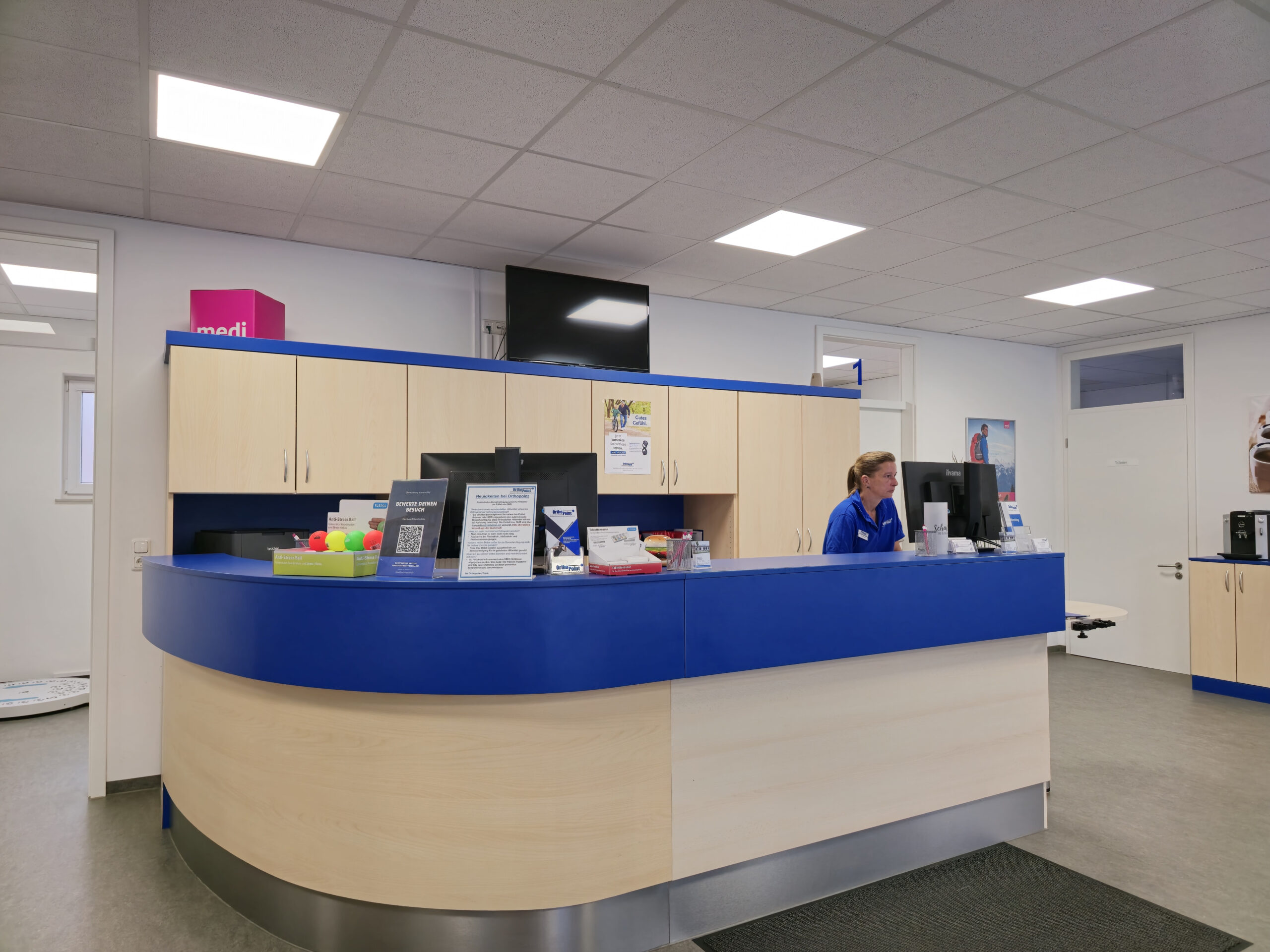 Ladenzeile in einem Sanitätshaus mit blauen Akzenten und einer Fachverkäuferin von Orthopoint die an einem PC arbeitet