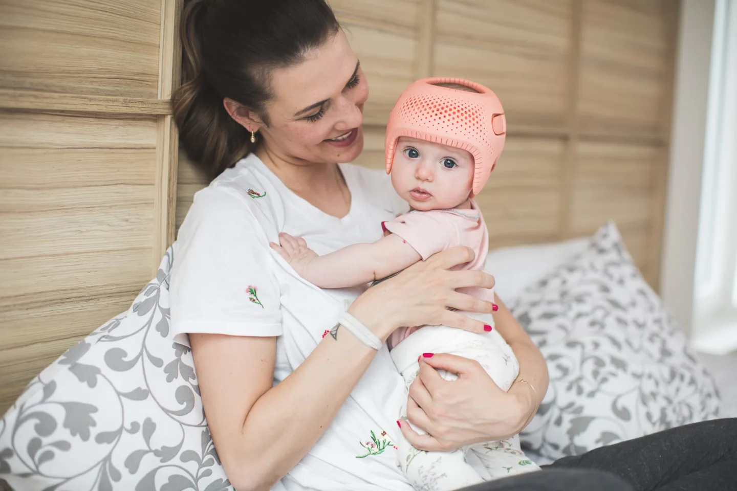 Einen Mama wie sie strahlend ihr Baby in den Händen hält. Das Baby trägt eine rosane Kopforthese.