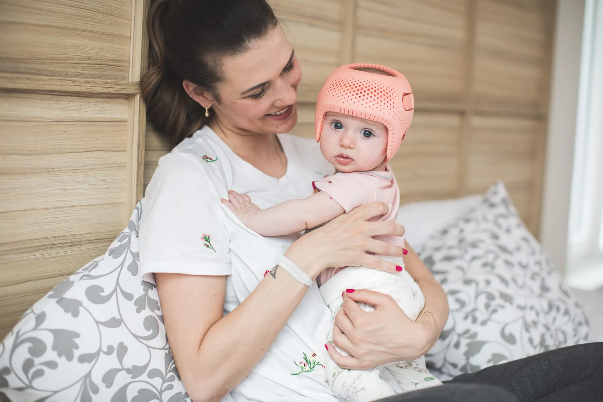 Einen Mama wie sie strahlend ihr Baby in den Händen hält. Das Baby trägt eine rosane Kopforthese.